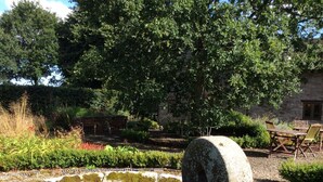 Ein steinerner Brunnen mit Holzbalken, umgeben von einem Garten mit verschiedenen Pflanzen und einem Steinhaus im Hintergrund.