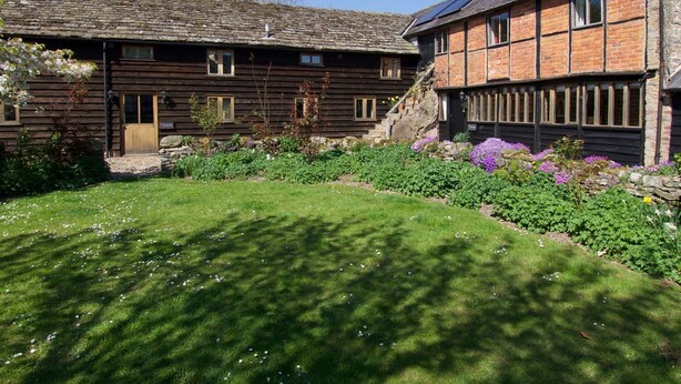 Ein traditionelles Holzhaus mit steinernen Fundament, ein gepflegter Garten mit verschiedenen Pflanzen und Blumen sowie ein klarer, blauer Himmel.