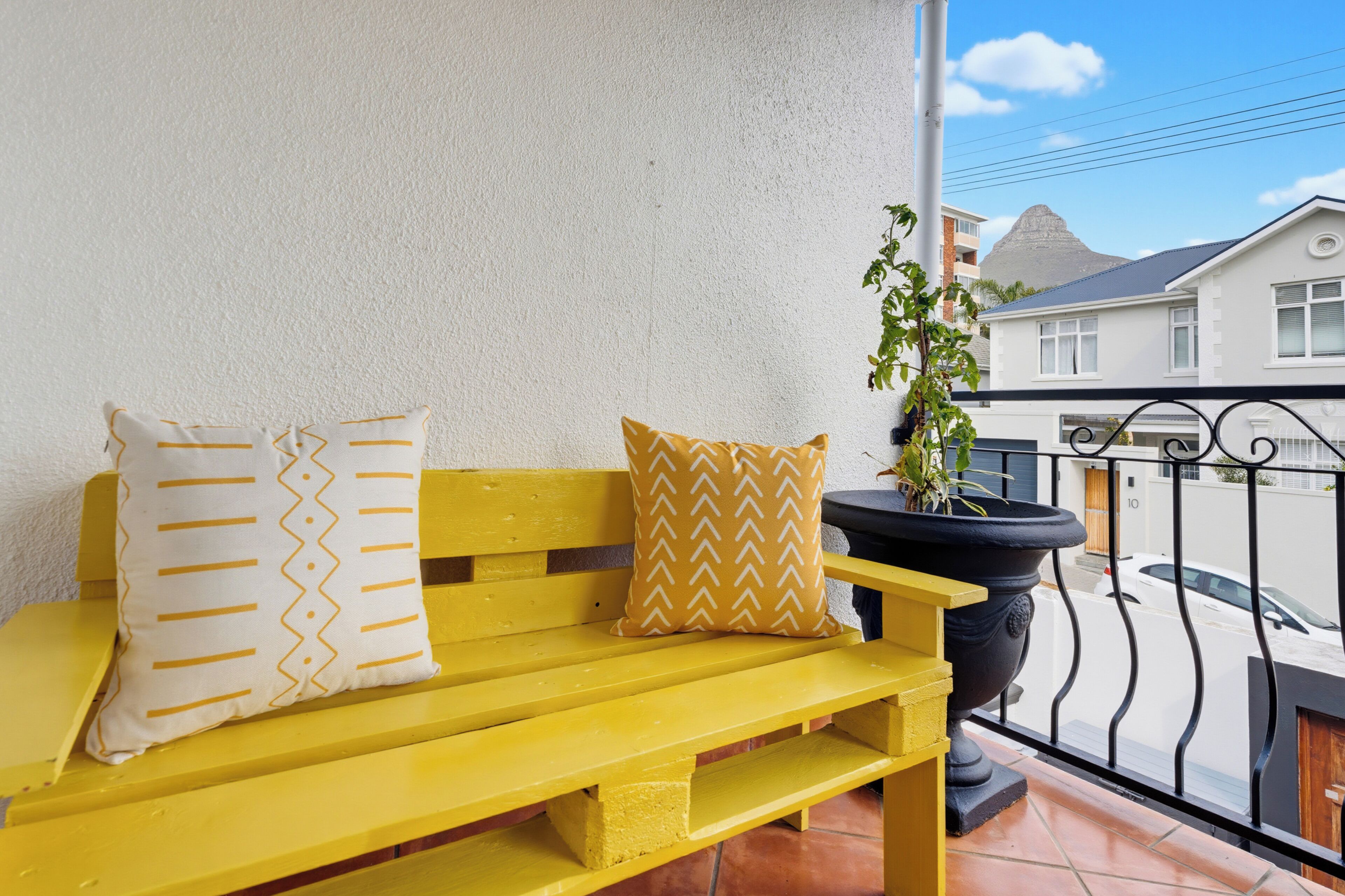 A yellow bench with patterned cushions, a potted plant, and a view of a residential area with a house and a street.