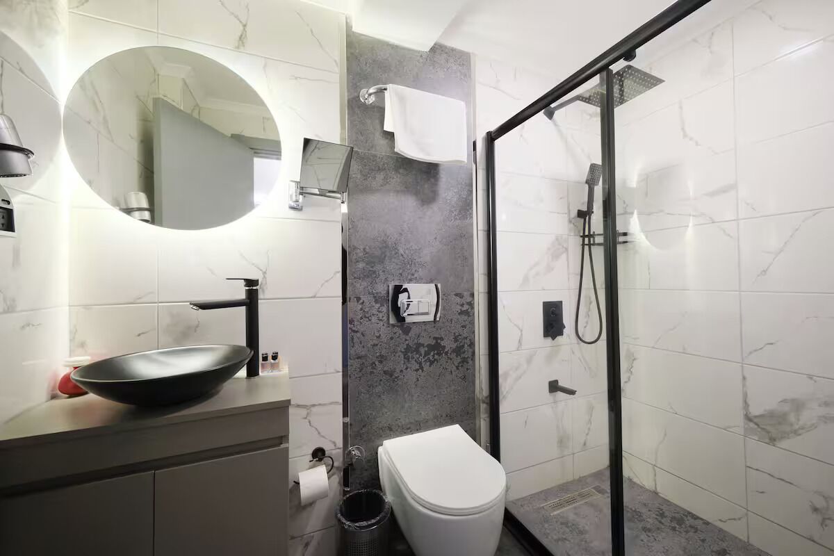 Un baño moderno con un espejo redondo, un lavabo negro, un inodoro y una zona de ducha con mampara de vidrio.