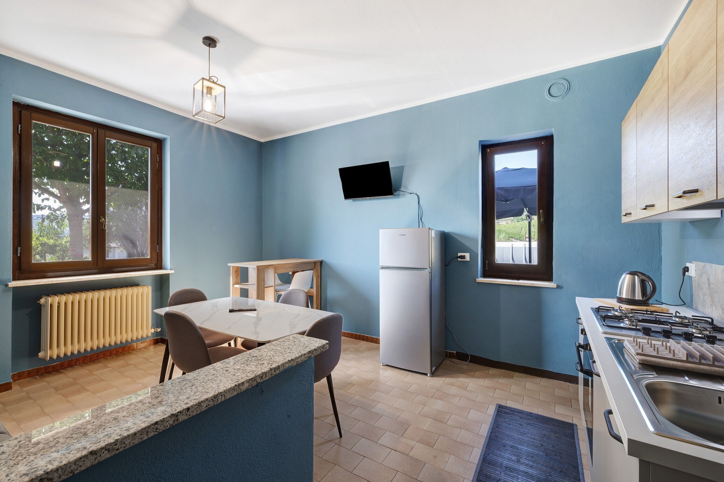 A compact kitchen with a built-in refrigerator, a dining table with chairs, a TV mounted on the wall, and a window with a view of the outdoors.