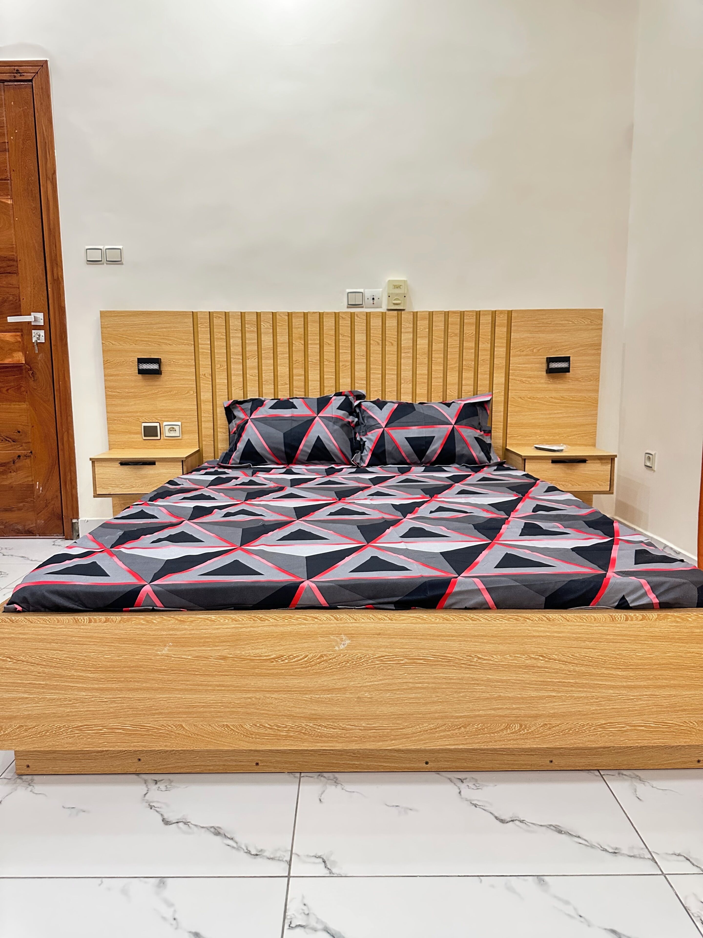 A bed with a geometric patterned bedspread, wooden headboard, bedside tables, and a marble-tiled floor.