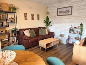 Ein gemütliches Wohnzimmer mit einem braunen Ledersofa, einem Holztisch und Armstühlen in Türkis. Ein Bücherregal beherbergt dekorative Gegenstände, und eine an der Wand befestigte Schilder zeigt den Schriftzug 'love you to the beach'.