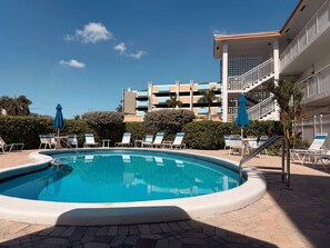 Ein Schwimmbecken mit Liegestühlen und Sonnenschirmen, umgeben von einem Gebäude mit mehreren Balkonen und blauem Himmel.
