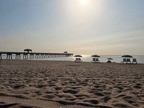 Ein Strand mit einem Steg, der ins Wasser ragt, mehreren Strandsonnenschirmen und Liegestühlen.