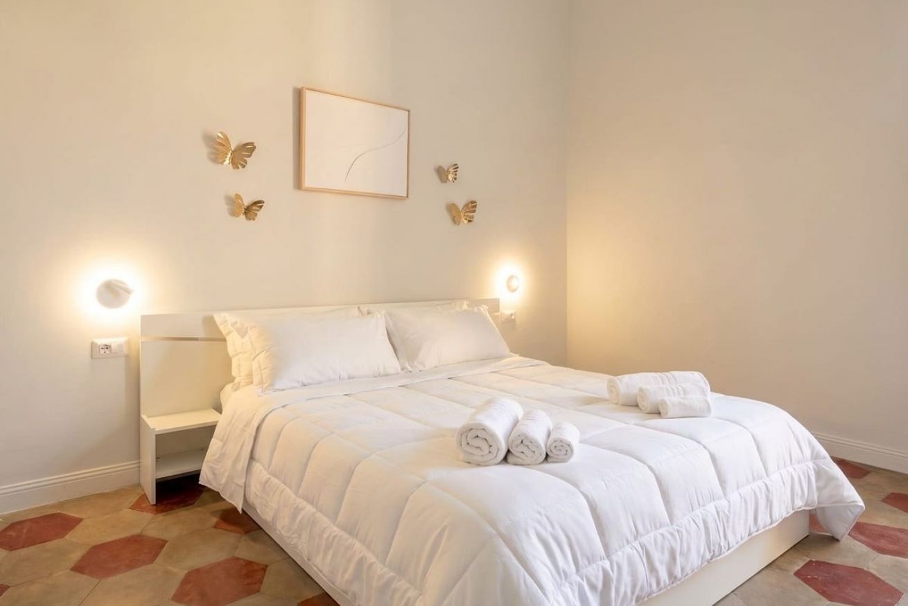 A neatly made bed with white bedding and rolled towels, a bedside table, and wall-mounted light fixtures.
