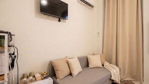 Ein Zimmer mit grauem Sofa, beigen Kissen, einem Flachbildfernseher, einer Klimaanlage, einem Vorhang und einem kleinen Tisch mit Gegenständen darauf.