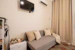 A room with a grey sofa, beige cushions, a flat-screen TV, an air conditioner, a curtain, and a small table with items on it. - City Hub 1 | Boutique Studio in Bari's Historic Heart (Bari)
