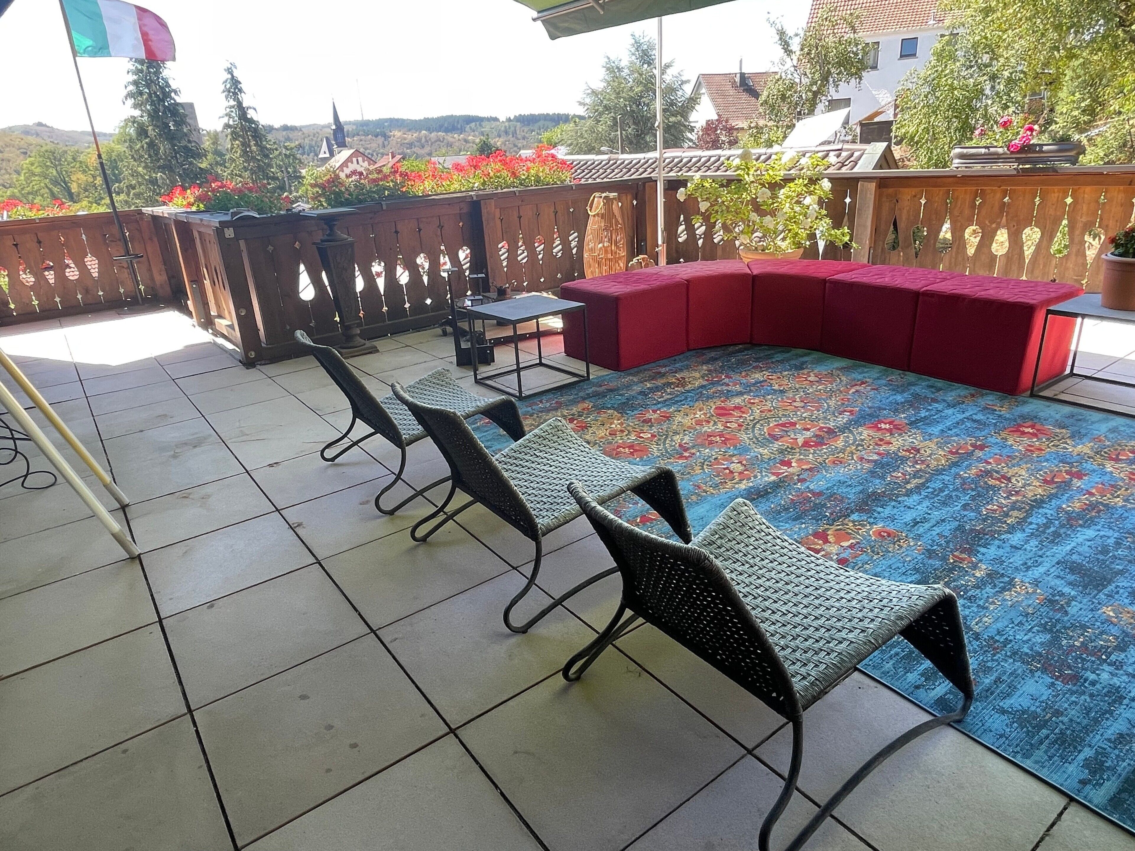 Een terras met betegelde vloer, een rode, beklede bank en twee rieten stoelen.