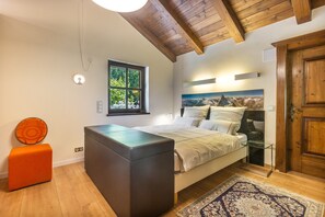 A bedroom with a bed, a window, a door, and a small orange ottoman.