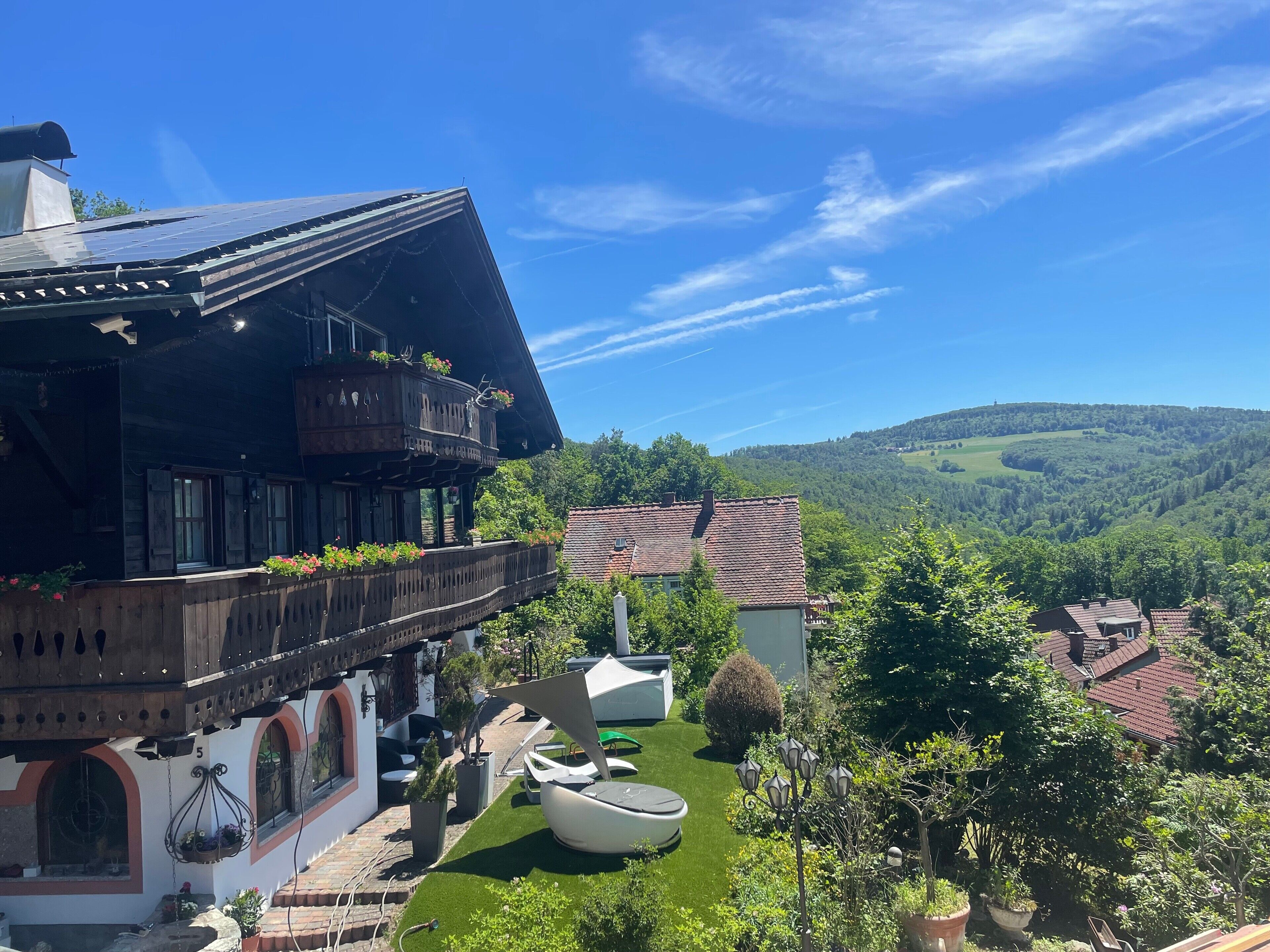Une maison traditionnelle en bois, dotée d’un balcon, d’un petit jardin avec un bateau blanc, et offrant une vue sur un paysage vallonné.