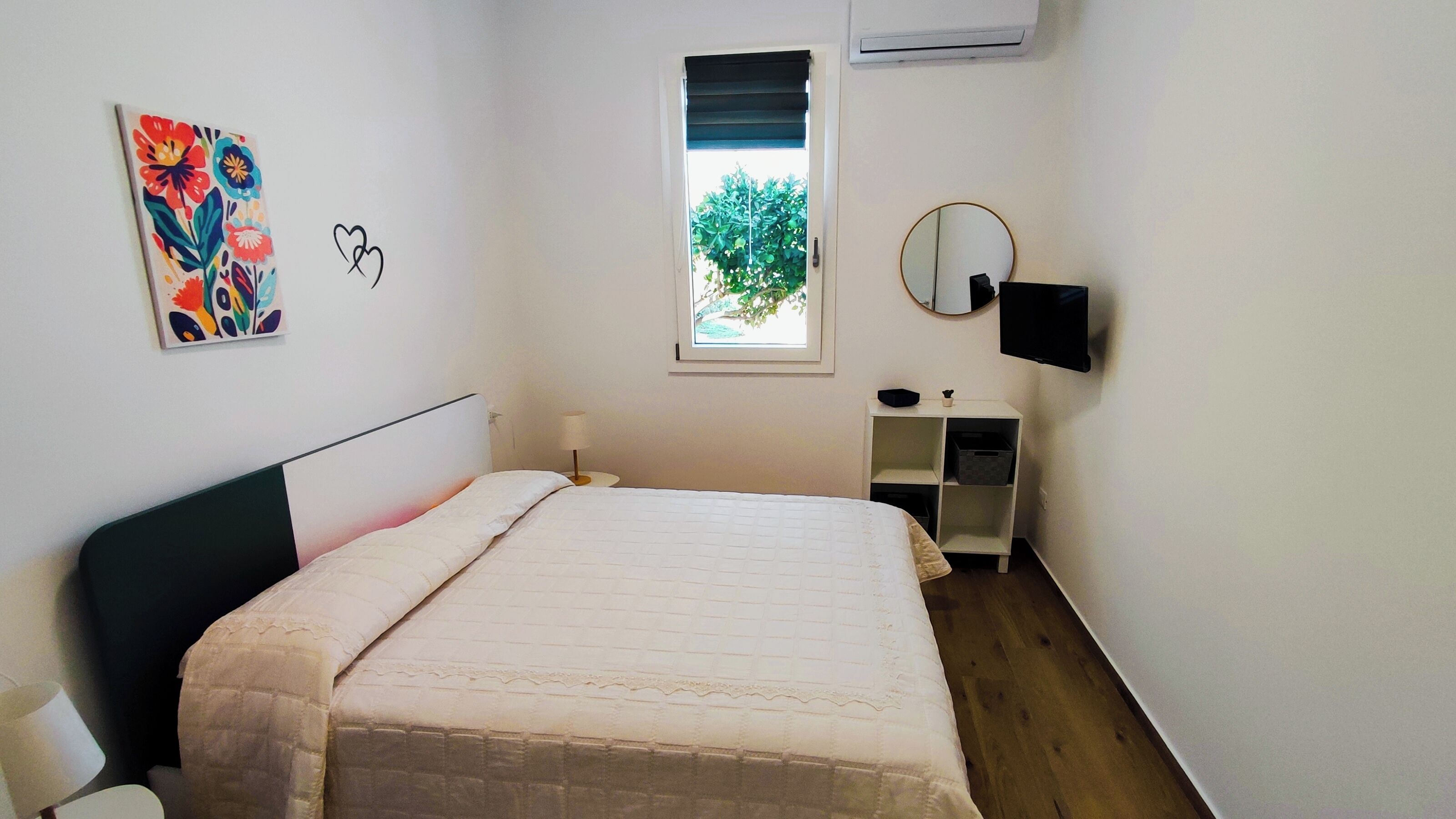 A bedroom with a bed, a bedside table, a mirror, a TV, a window, and a painting on the wall.