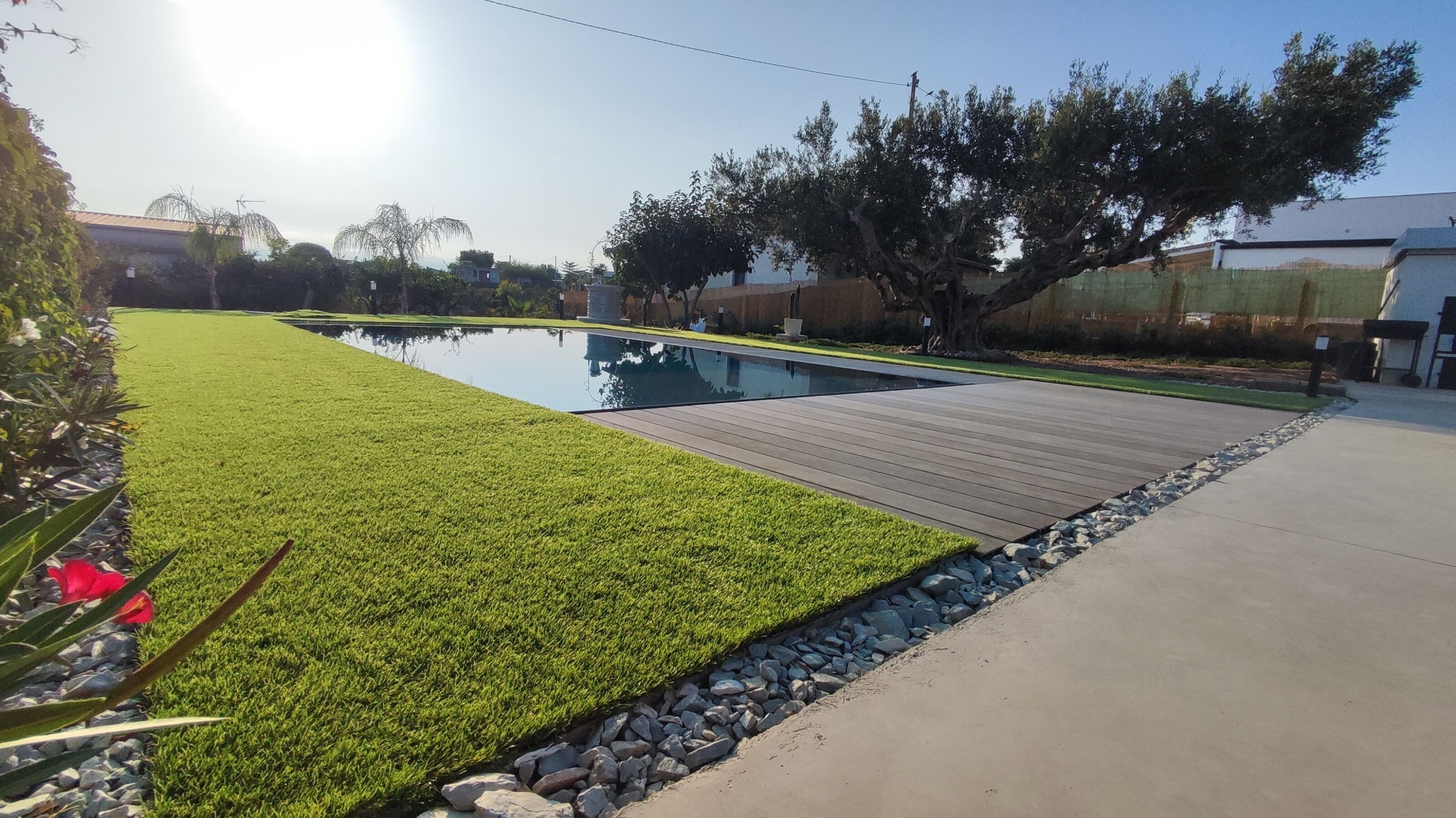 A swimming pool with a wooden deck, surrounded by a well-maintained lawn and a stone border.