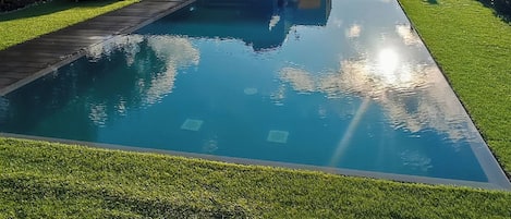 Una piscina con superficie de agua azul cristalina, rodeada por un césped cuidado y un edificio con techo azul.
