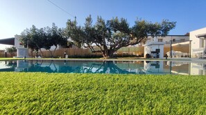 A swimming pool with a grassy foreground, trees, and a building with outdoor seating. - Villa du Cori: Pool and Relaxation (Alcamo)