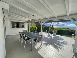 Eine überdachte Terrasse mit Esstisch und Stühlen, einer hängenden Leuchte und Blick aufs Meer.