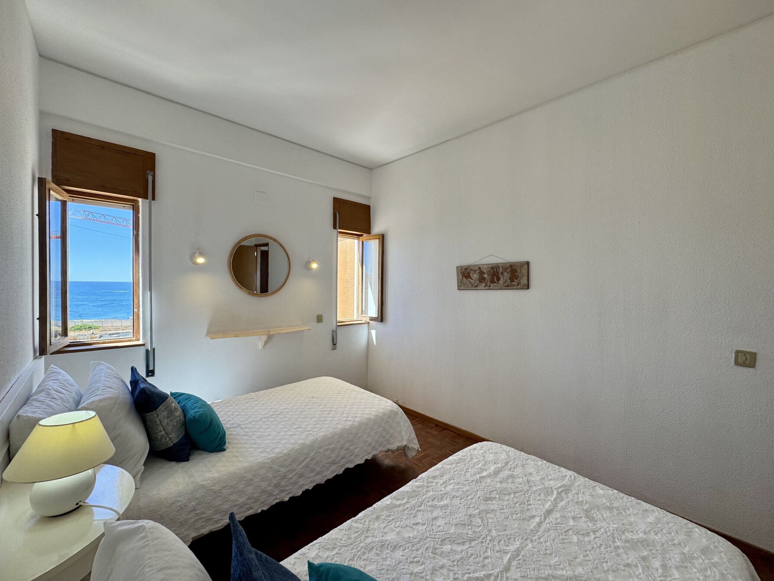 Quarto com duas camas, uma mesa de cabeceira com abajur, um espelho e janela com vista para o mar.