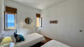 Quarto com duas camas, uma mesa de cabeceira com abajur, um espelho e janela com vista para o mar.