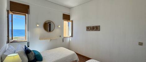 A bedroom with two beds, a bedside table with a lamp, a mirror, and a window with a view of the sea.