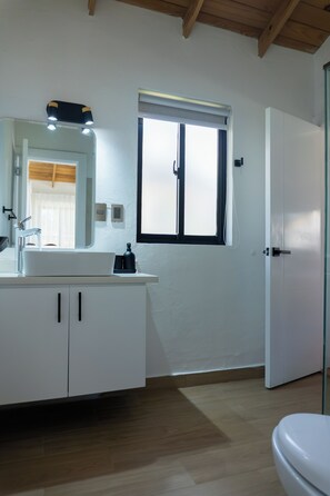 Un baño con lavamanos blanco, espejo y armarios, una ventana con persianas y una puerta.