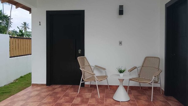 Uma varanda coberta com duas cadeiras de vime, uma mesa pequena e um vaso de planta.