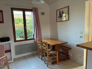 A room with a wooden table and chairs, a window with curtains, a radiator, and a framed picture on the wall. - Detached 2-room house with a garden, within walking distance of the beach and shops! (Saint-Lunaire)
