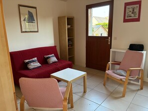 A living room with a red sofa, two striped chairs, a white table, a wooden bookshelf, and a door leading outside. - Detached 2-room house with a garden, within walking distance of the beach and shops! (Saint-Lunaire)