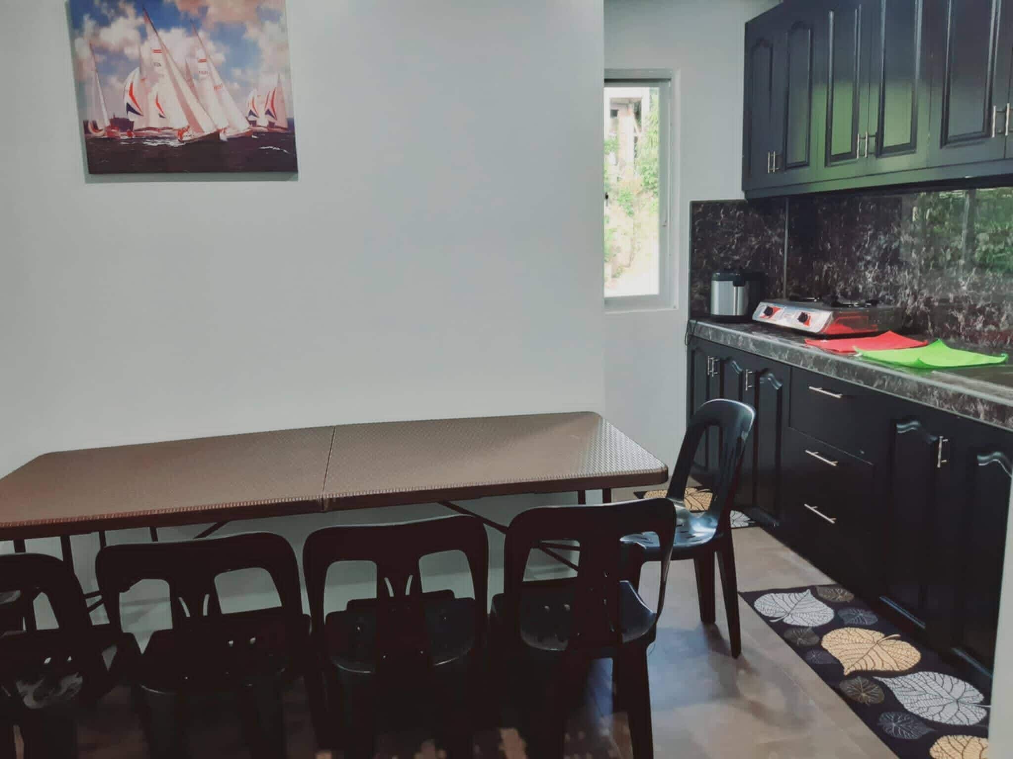 A kitchen with a dining table and chairs, a painting of sailboats, and a view of greenery outside.