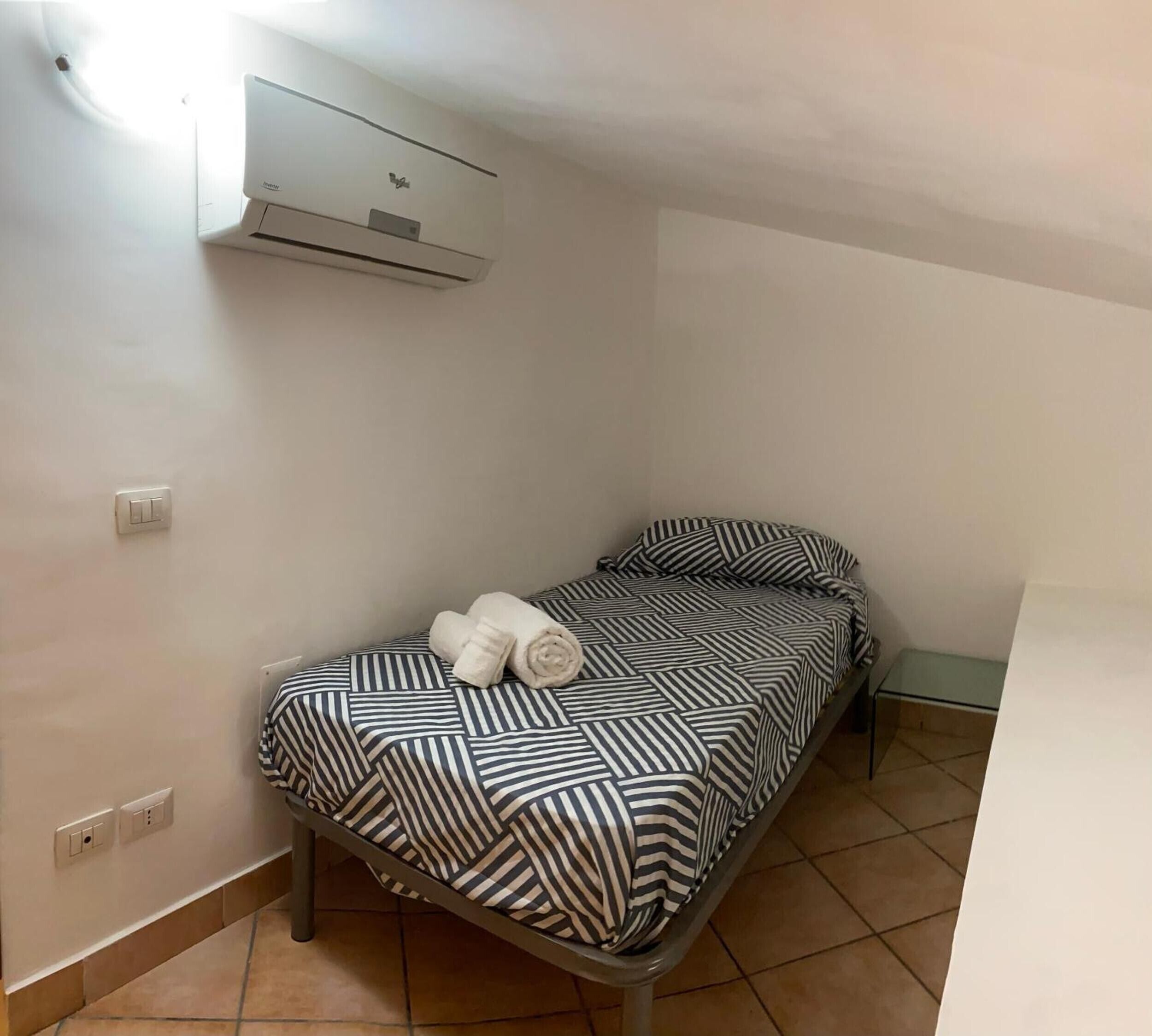 A small, single-bed room with a patterned bedspread, a rolled towel on the bed, an air conditioning unit mounted on the wall, and a small table with a glass top.