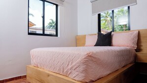 Uma cama de solteiro com estrutura de madeira e colcha rosa acolchoada em quarto com paredes brancas e janela vista para palmeiras.