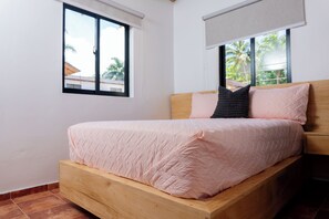 Ein Einzelbett mit Holzrahmen und einer rosa gesteppten Bettdecke in einem Zimmer mit weißen Wänden und einem Fenster mit Blick auf Palmen.