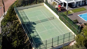 Ein Außen-Tennisplatz mit umgebendem Zaun, neben einem Gebäude mit Schwimmbad.