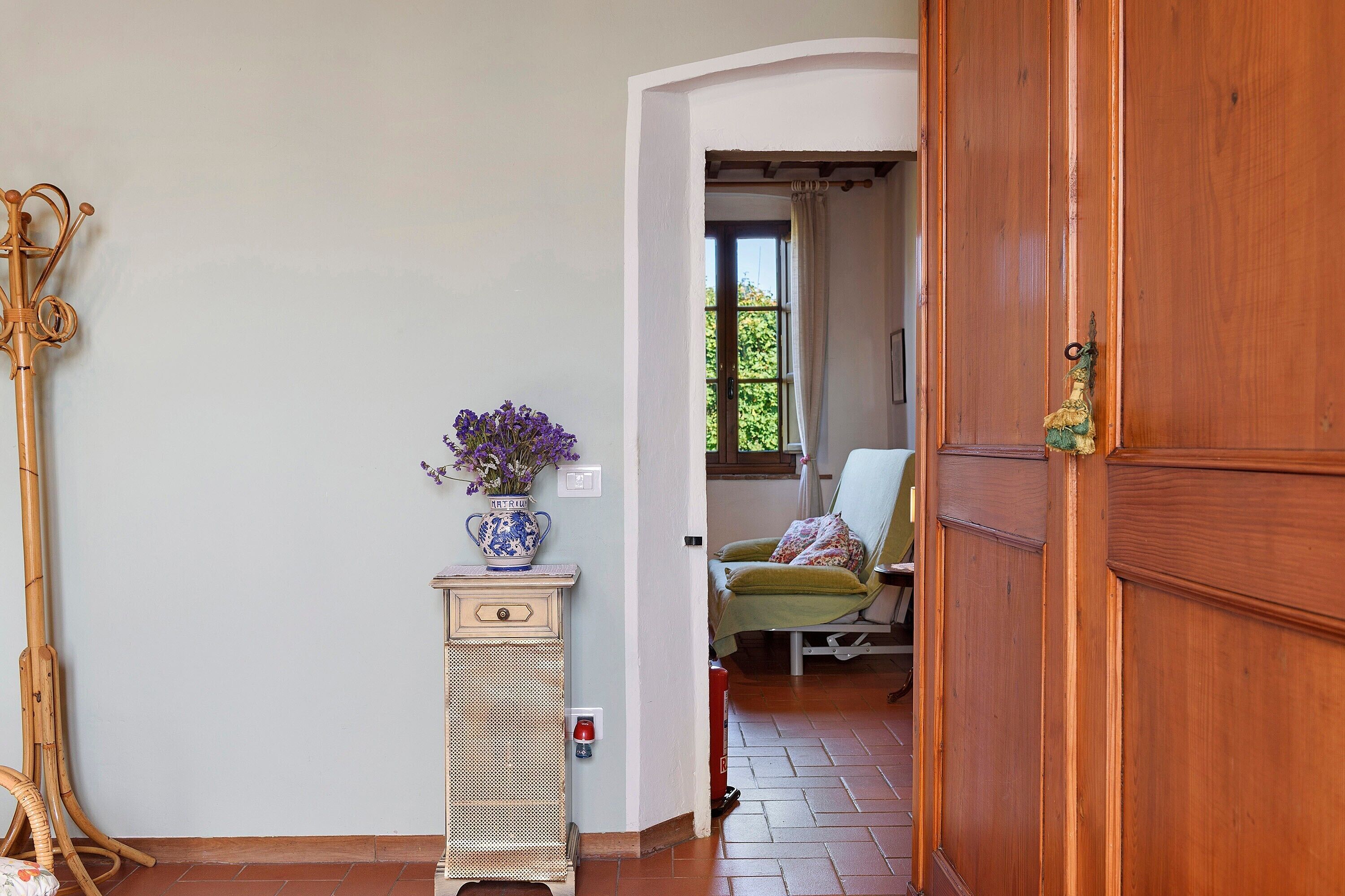 A room with a wooden door, a wicker chair, a small table with a vase of flowers, and a view through an open door to another room.