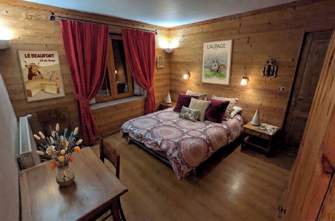 A cozy wooden room with a bed, a nightstand, a table with a vase, and a window with curtains.