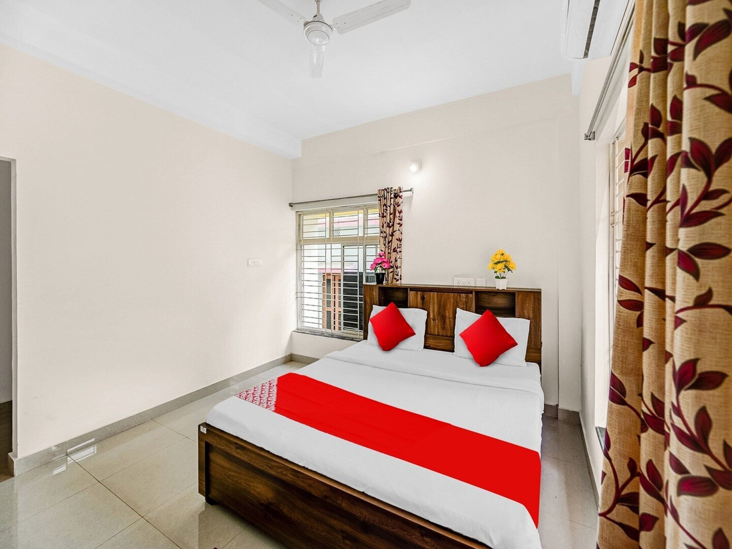 A bedroom with a bed, a window with curtains, a ceiling fan, and a bedside table with a vase of flowers.