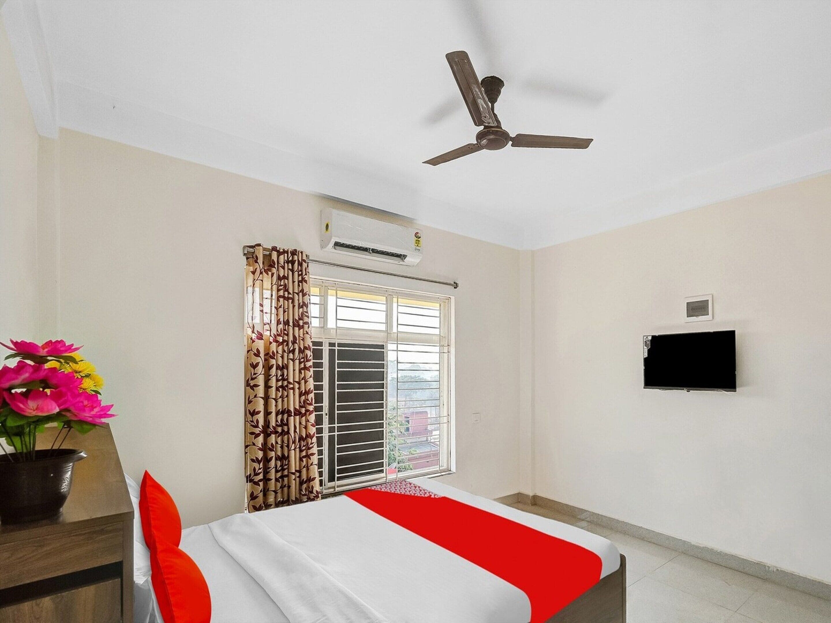 A hotel room with a bed, a ceiling fan, an air conditioning unit, a window with curtains, a television, and a potted plant.