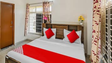 Une chambre d’hôtel avec un lit, une tête de lit en bois, une fenêtre avec des rideaux, une porte et une petite table sur laquelle est posé un vase de fleurs.