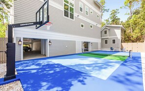 A basketball court with a Dominator brand hoop, a covered parking area, and a house with multiple windows and a balcony. - Private 30A Resort: Infinity Pool, Gym, Pickleball, 2 min. to Beach. Must see! (Santa Rosa Beach)