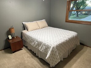 A bedroom with a bed, a nightstand, a lamp, and a window with a view of a tree and a house.