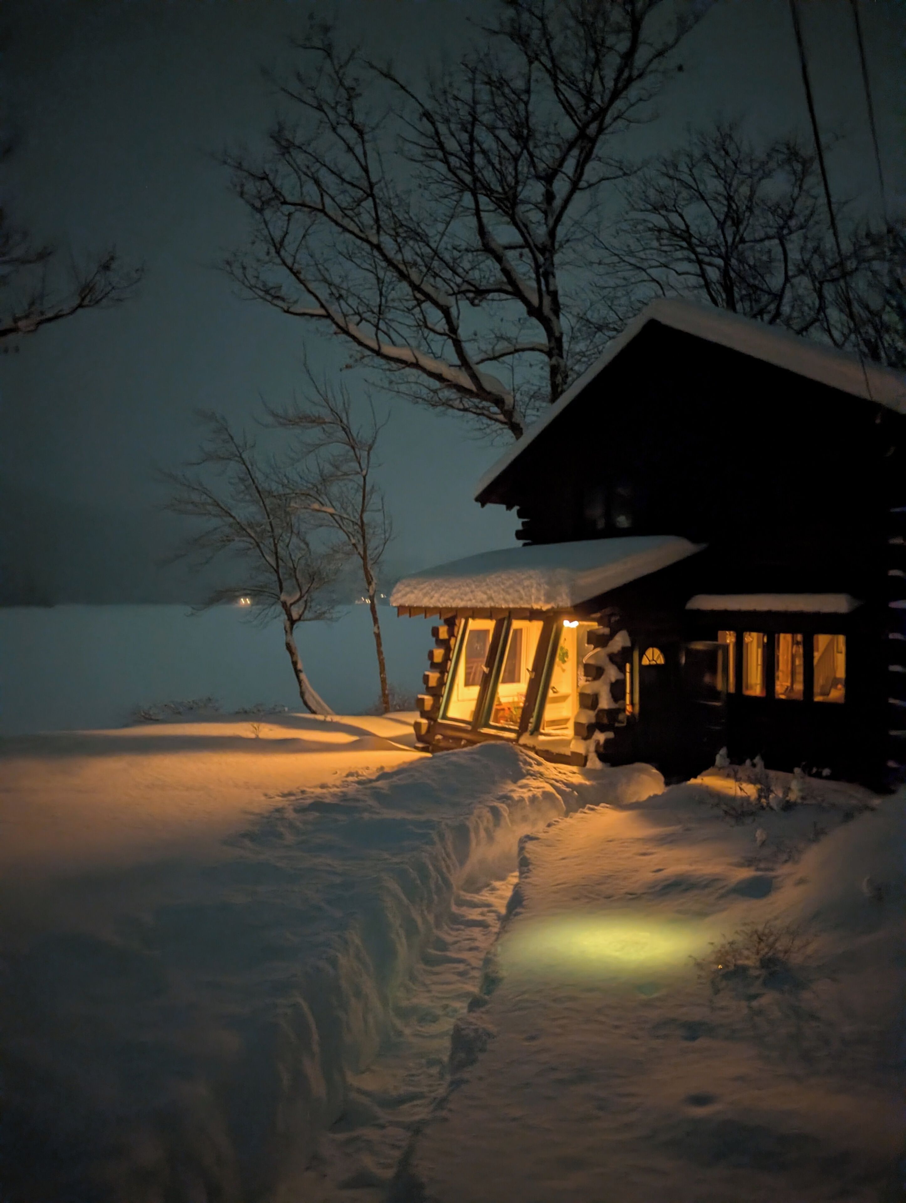 Una cabaña de troncos con techo nevado e interior iluminado, rodeada de nieve y árboles desnudos.