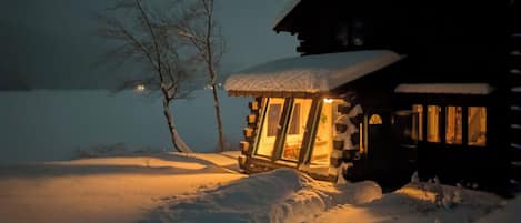 Une cabane en rondins, dont le toit est recouvert de neige et dont l’intérieur est éclairé, entourée de neige et d’arbres sans feuilles.