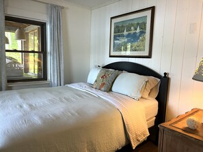 Ein Bett mit Kopfteil, ein Fenster mit Vorhängen, ein gerahmtes Bild und ein Nachttisch mit Lampe.