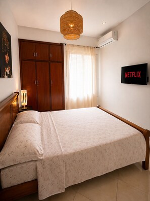 Ein Schlafzimmer mit Bett, Kleiderschrank, Nachttischlampe, Fenster mit Vorhängen, Klimaanlage und einem an der Wand befestigten Fernseher, der das Netflix-Logo zeigt.