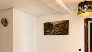 A hallway with a red patterned rug, two green ottomans, a painting of a waterfall, and a ceiling light fixture. - Family Apartment for 4–5 People (Fes)