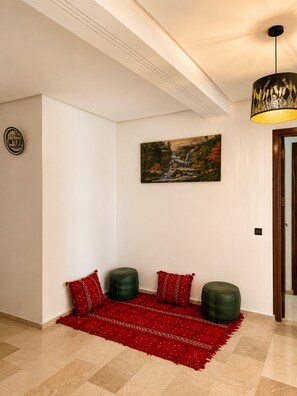 A hallway with a red patterned rug, two green ottomans, a painting of a waterfall, and a ceiling light fixture. - Family Apartment for 4–5 People (Fes)