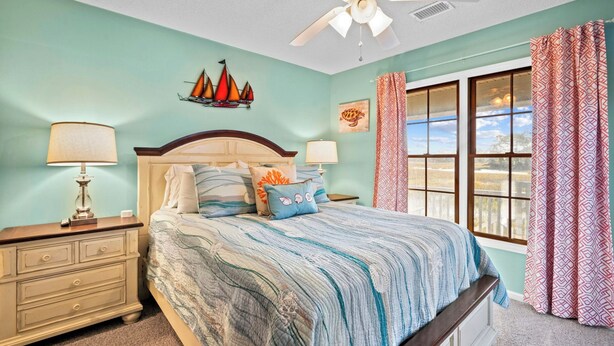 A bedroom with a bed, two bedside tables, a ceiling fan, a window with curtains, and a wall-mounted ship decoration. - Beautiful Marsh Views, Golf Cart Included, Elevator - 364 Blue Gill (Fripp Island)