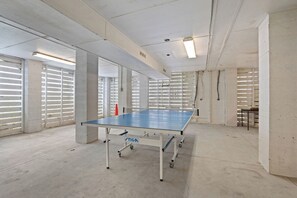 A ping pong table in a spacious, industrial-style room with concrete walls and ceiling, large windows with blinds, and a visible ventilation system. - Walk to the Beach, Elevator, Golf Cart Included - 619 Dolphin Rd (Fripp Island)