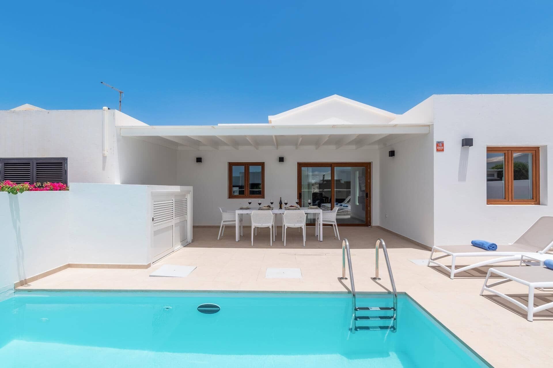 Un espace piscine avec une table et des chaises, un coin salon abrité et un ciel d’un bleu limpide.
