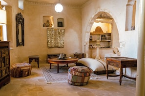 A traditional room with a round wooden table, a sofa, a patterned ottoman, a mirror, and a bookshelf.