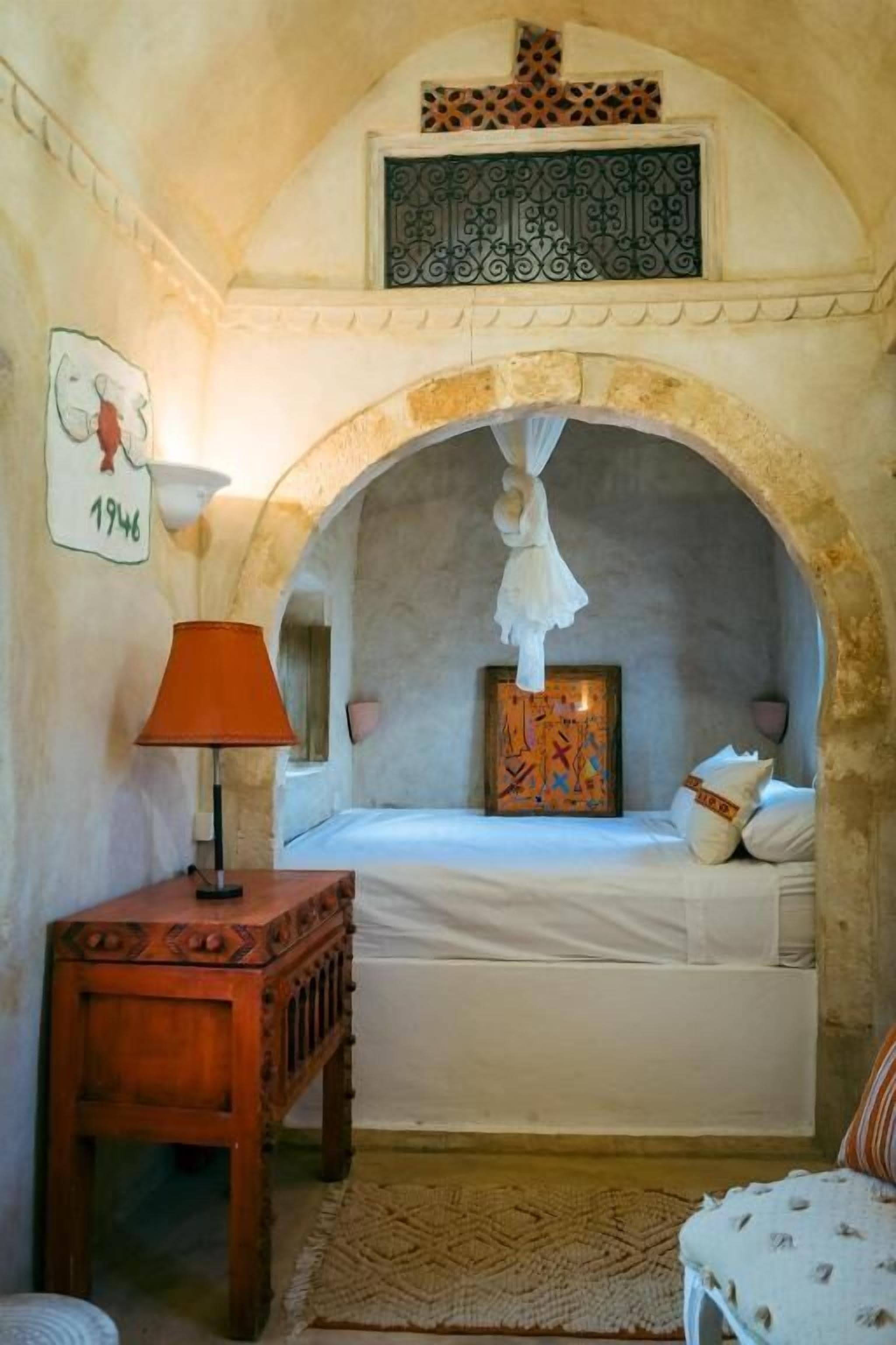 A bedroom with a bed under an arched alcove, a bedside table with a lamp, and a framed picture on the wall.